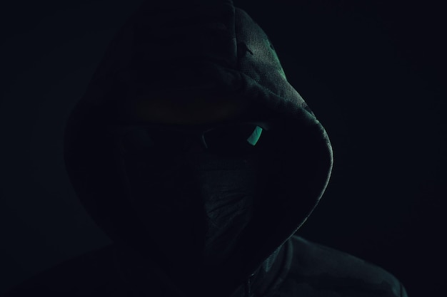 Foto stylischer mann, dunkler hoodie, schwarze brille und maske, grüner hintergrund, hacker-angreifer und bandit