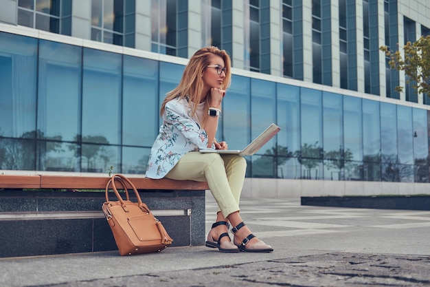 Stylischer Modeblogger entspannt sich im Freien, arbeitet am Laptop und sitzt auf einer Bank vor einem Wolkenkratzer.