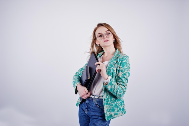 Stylisches blondes Mädchen in Jacke und Jeans mit Brille mit Notizbuch, Tagebuch und Laptop in der Hand vor weißem Hintergrund im Studio