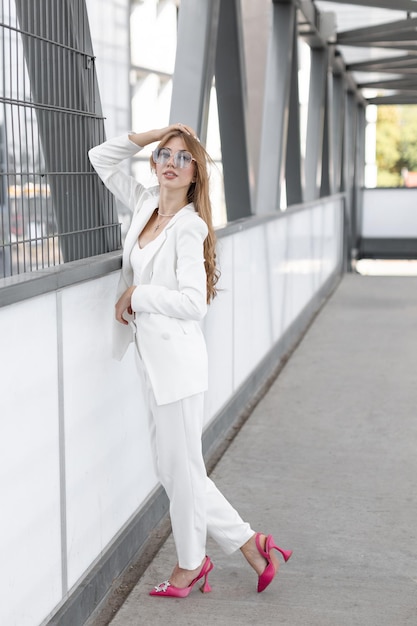 Stylish adorável mulher bonita com cabelos ondulados vestindo fato branco elegante e óculos de sol de pé e se divertindo na rua