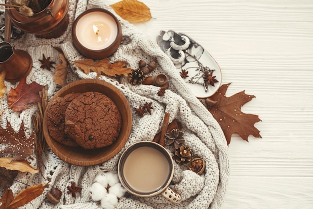 Stylish Autumn Flat Lay Köstliche Kaffeekekse und schöne Herbstblätter Kerze Nüsse Eicheln Baumwolle Zimt auf Pullover und weißer rustikaler Hintergrund Draufsicht Gemütliches Herbstbild