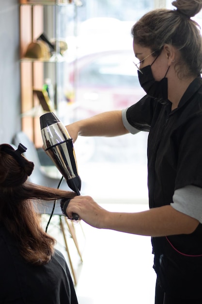 Stylistin, die in einem Salon mit viel natürlichem Licht einen Fön und einen runden Kamm durch die Haare ihrer Kundin führt