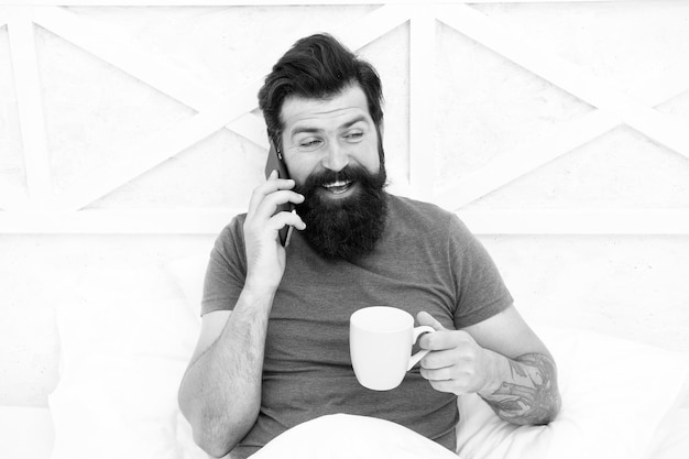 Sua rotina matinal Homem barbudo bebe café da manhã na cama Hipster fala no celular pela manhã Sua xícara matinal de bom humor Vida moderna Nova tecnologia Estilo de vida móvel
