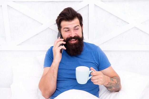 Sua rotina matinal Homem barbudo bebe café da manhã na cama Hipster fala no celular pela manhã Sua xícara matinal de bom humor Vida moderna Nova tecnologia Estilo de vida móvel