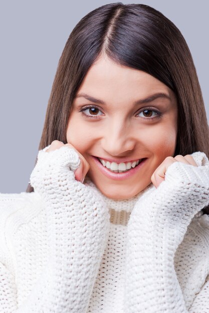 Suavidad y calidez. Primer plano de mujeres jóvenes en suéter blanco tomados de la mano cerca de la cara y sonriendo mientras está de pie contra el fondo gris