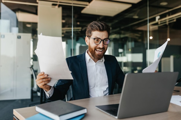 Sucesso nos negócios Empresário muito feliz agitando os punhos segurando documentos depois de muito olhar para a tela do laptop