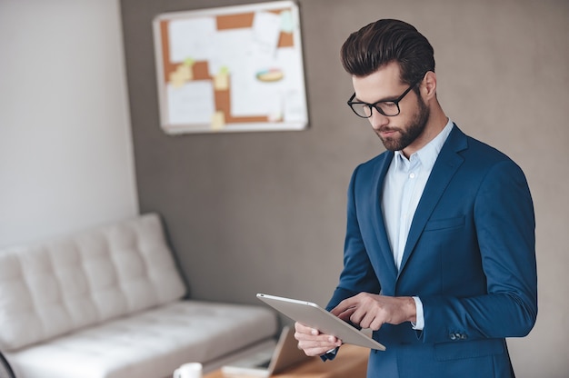 Suche nach Lösung. Hübscher junger Mann mit Brille und Arbeiten mit Touchpad im Stehen im Büro