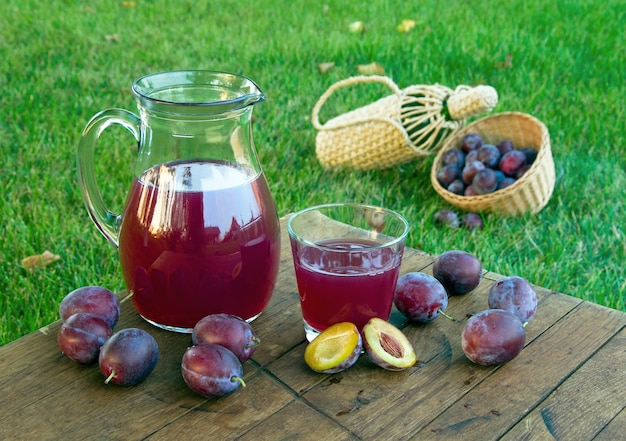 Suco de ameixa na garrafa e vidro com ameixas em um fundo de gramado verde. Tiro horizontal ao ar livre.