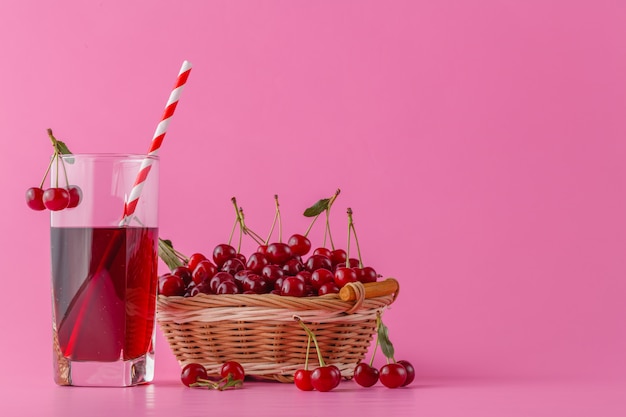 Suco de cereja em um copo e jarra em rosa com frutos maduros na cesta de vime