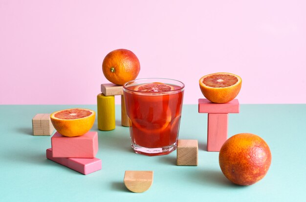 Suco de laranja em um copo com podims de formas geométricas e laranjas em fundo de hortelã e rosa