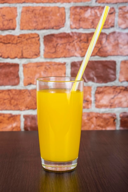 Foto suco de laranja em um copo com um tubo sobre uma mesa de madeira e um fundo de parede de tijolos