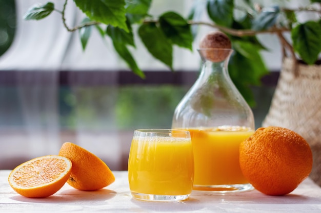 Suco de laranja fresco em jarro e vidro com laranjas maduras na mesa de madeira branca no terraço de verão