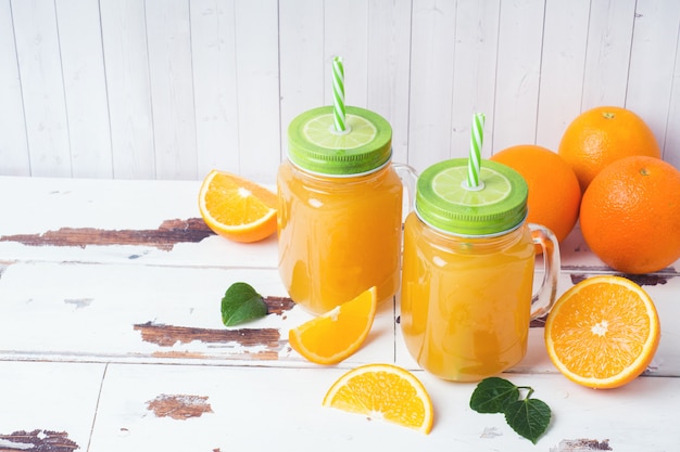 Suco de laranja nos frascos de vidro e laranjas frescas em um fundo rústico de madeira branco.