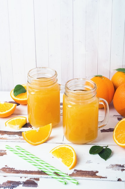 Suco de laranja nos frascos de vidro e laranjas frescas em um fundo rústico de madeira branco.