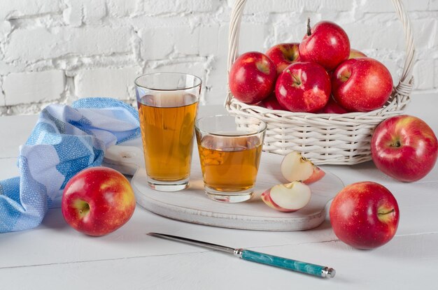 Suco de maçã em dois copos, uma cesta com maçãs vermelhas e um guardanapo azul contra uma parede branca.