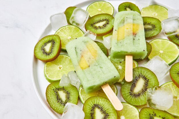 Suco natural orgânico congelado no palito com uma fatia de manga em uma tigela com pedaços de limão, kiwi e cubos de gelo em uma mesa de mármore cinza com uma cópia do espaço para texto. Vista do topo