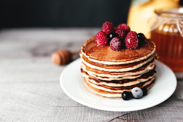 Suculentas panquecas com frutas e mel em um prato branco, colher, pote, mesa de madeira, vaso amarelo com eucalipto. Foto de alta qualidade