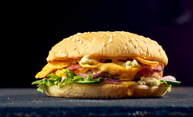 Suculento hambúrguer com carne de porco, queijo, ovo, pepino e tomate, molho em um fundo escuro.