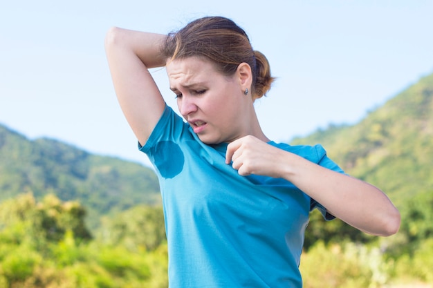 Foto sudorosa niña olisqueando su axila, mirando el lugar, sudor manchado en su camiseta con disgusto emoción, cara, frunce el ceño. mujer frustrada molesta que sufre de hiperhidrosis. día soleado de verano