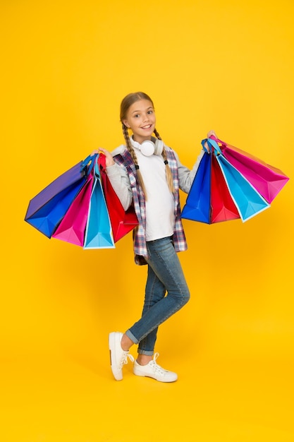 Süchtiger Verbraucher Tauchen Sie ein ins Einkaufen Glückliches Kind mit Papiertüten Kleines Mädchen lächelt mit Einkaufstüten auf gelbem Hintergrund Urlaubsvorbereitung und Feier Einkaufen und Verkauf am schwarzen Freitag