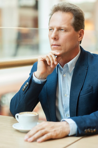 Sueños empresariales. Hombre maduro pensativo en ropa formal tomando café y sosteniendo la mano en la barbilla mientras está sentado en la cafetería.