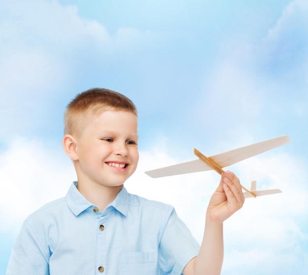 sueños, futuro, hobby y concepto de infancia - niño sonriente sosteniendo un modelo de avión de madera en su mano sobre fondo de cielo azul