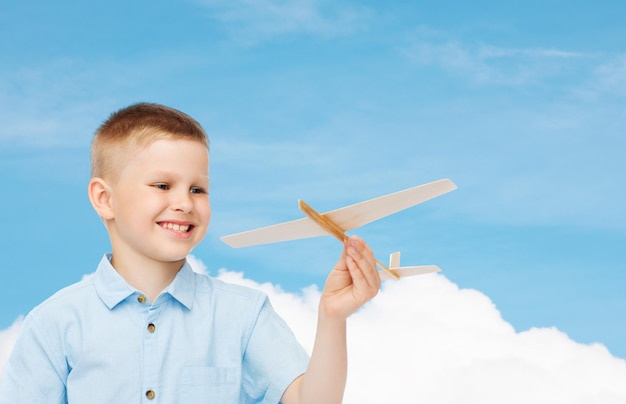 sueños, futuro, hobby y concepto de infancia - niño sonriente sosteniendo un modelo de avión de madera en su mano sobre fondo de cielo azul