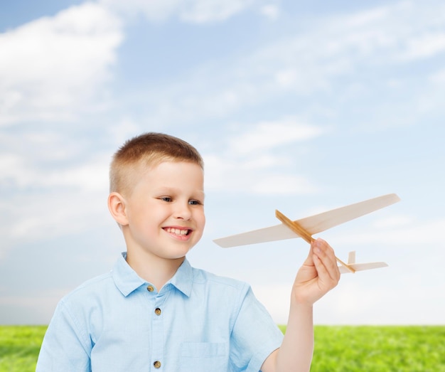 sueños, futuro, hobby, naturaleza y concepto de infancia - niño pequeño sonriente sosteniendo un modelo de avión de madera en su mano sobre fondo natural