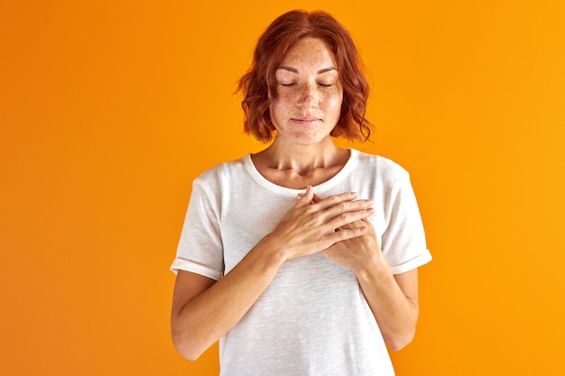 Sueños de mujer con los ojos cerrados, tomados de la mano en el pecho aislado en estudio sobre fondo naranja