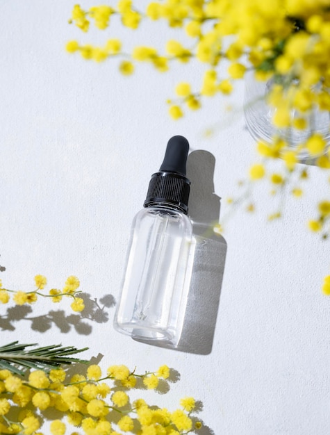 Suero facial y corporal en botella sobre un fondo claro con flores de mimosa y sombra dura Salud y belleza