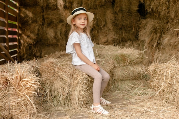 Süß lächelndes kleines Mädchen mit Weidenhut, das an einem sonnigen Sommertag auf Heuhaufen im Heuboden sitzt Glückliche Kindheit und ländliches Urlaubskonzept