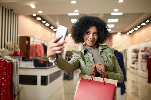 Süße afrikanisch-amerikanische Frau macht Selfie mit Einkaufstüten und lächelt in der Nähe eines Bekleidungsgeschäfts schwarz