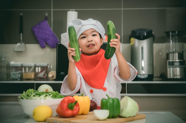 Süße asiatische Mädchen tragen Kochuniform mit viel Gemüse auf dem Tisch im KüchenraumMachen Sie Essen zum AbendessenLustige Zeit für Kinder