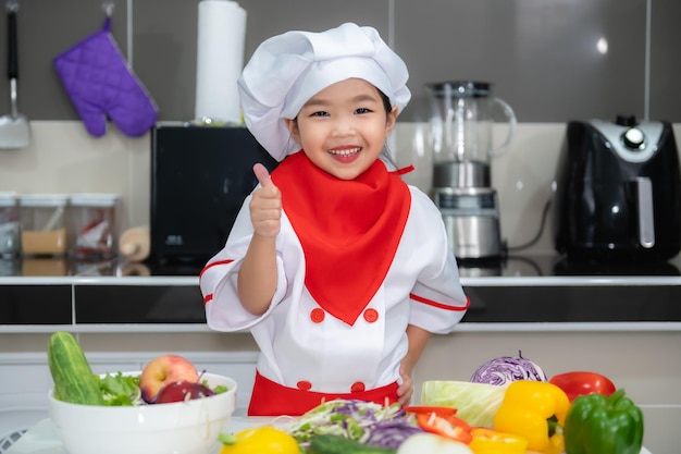 Süße asiatische Mädchen tragen Kochuniform mit viel Gemüse auf dem Tisch im KüchenraumMachen Sie Essen zum AbendessenLustige Zeit für Kinder