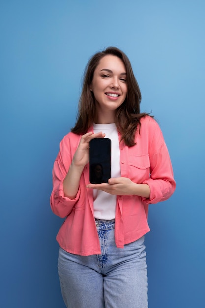Süße brünette Dame mit dunklem Haar unter den Schultern, Hemd und Jeans hält einen Smartphone-Bildschirm in der Hand