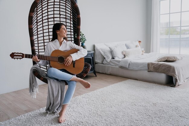 Süße Brünette in weißem Hemd und blauen Jeans sitzt in einem Korbsessel und spielt Gitarre