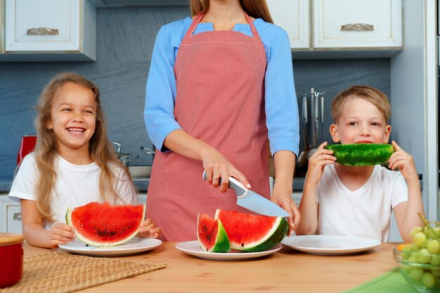 Süße Familie, Mutter und ihre Kinder essen Wassermelone in ihrer Küche und haben Spaß