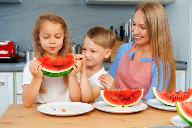 Süße Familie, Mutter und ihre Kinder essen Wassermelone in ihrer Küche und haben Spaß