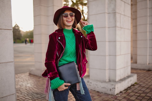 Süße Frau im trendigen Outfit im Herbststil, die auf der Straße mit lila Samtjacke, Sonnenbrille und Hut spazieren geht