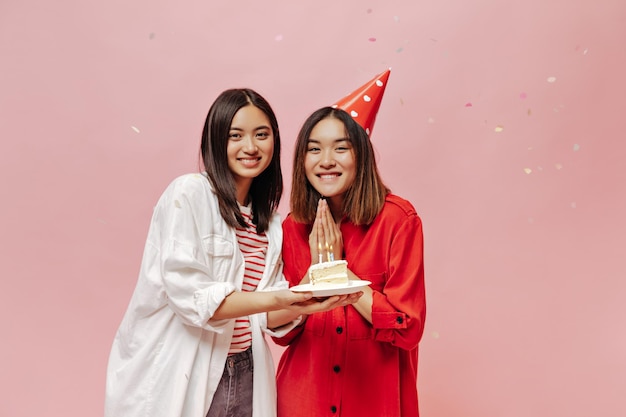 Süße fröhliche asiatische Mädchen feiern Geburtstag auf rosa Hintergrund mit Konfetti Junge Frau in weißem Hemd überreicht ihrer Freundin in Partyhut Bday-Kuchen