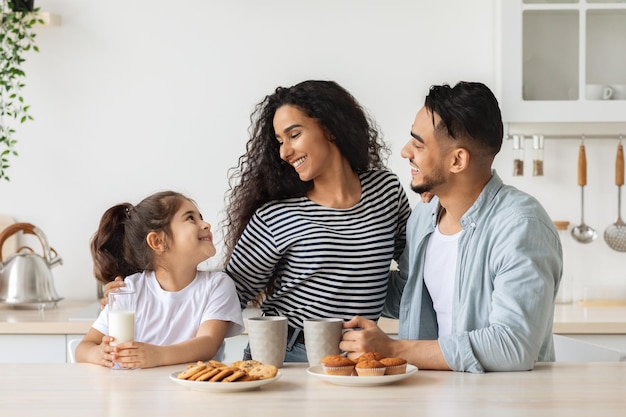 Süße, glückliche arabische Familie, die frühstückt, während sie das Wochenende zu Hause verbringt, fröhlicher junger Vater, Mutter und lockiges kleines Mädchen, die in der Küche sitzen, Kaffee und Milch mit hausgemachten Keksen trinken