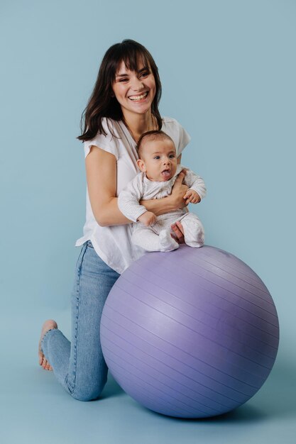 Süße junge brünette Mama macht Übungen mit Baby auf lila Gymnastikball auf blauem Hintergrund