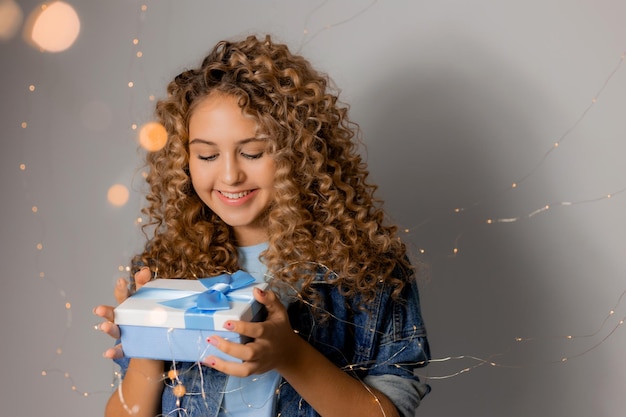 Süße junge Frau mit blauen Augen und blonden Locken in Denim-Kleidung hält eine Geschenkbox in ihren Händen. Mädchen öffnet das Geschenk. fester grauer Hintergrund. Platz für Text. Foto in hoher Qualität
