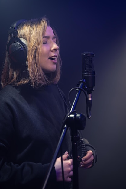 Süße junge Frau mit Kopfhörern singt in einem dunklen Raum in ein Mikrofon