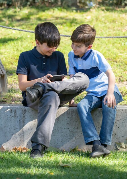 Süße junge Jungs sitzen im Park und spielen Online-Spiele am Telefon.