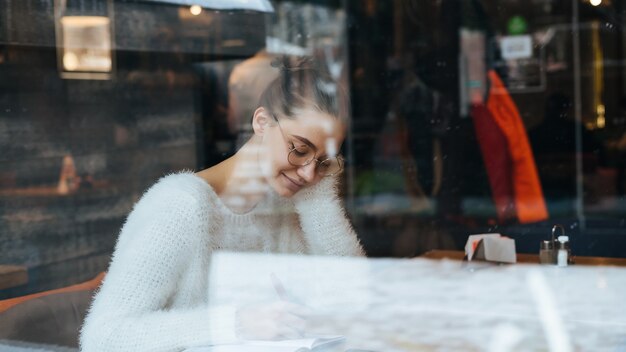 Süße junge Studentin mit Brille und einem weißen Pullover sitzt nach der Schule in einem Café, schreibt in ein Notizbuch