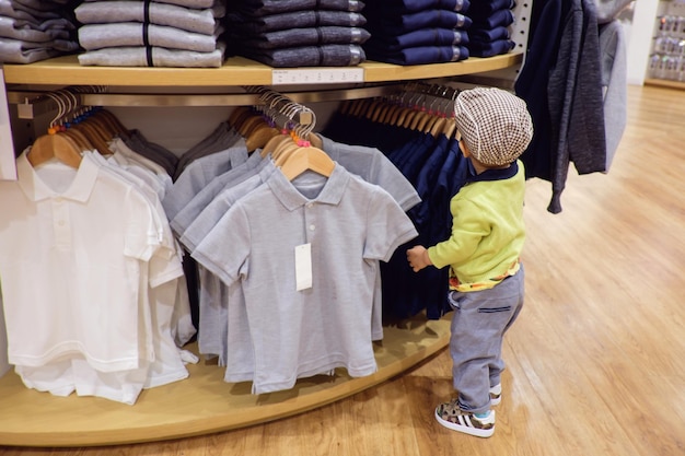 Süße kleine Asiatin, 18 Monate, 1 Jahr alt, Kleinkind, Junge, Kind, das sich für den Kauf von Kinderkleidung im Bekleidungsgeschäft entscheidet