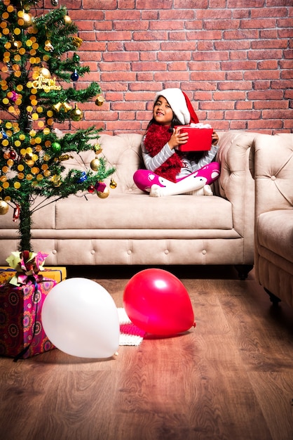 Süße kleine indische asiatische Kinder feiern Weihnachten zu Hause mit Weihnachtsmütze, Geschenken und Weihnachtsbaum