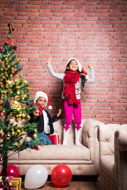 Süße kleine indische asiatische Kinder feiern Weihnachten zu Hause mit Weihnachtsmütze, Geschenken und Weihnachtsbaum