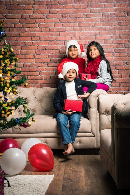 Süße kleine indische asiatische Kinder feiern Weihnachten zu Hause mit Weihnachtsmütze, Geschenken und Weihnachtsbaum